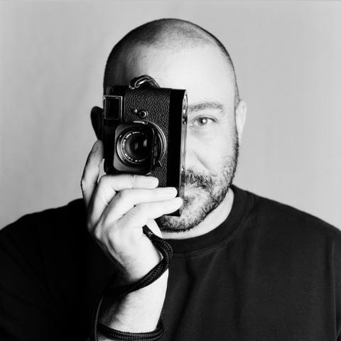 Stephen Vanasco holding a rangefinder camera to his eye, black-and-white headshot