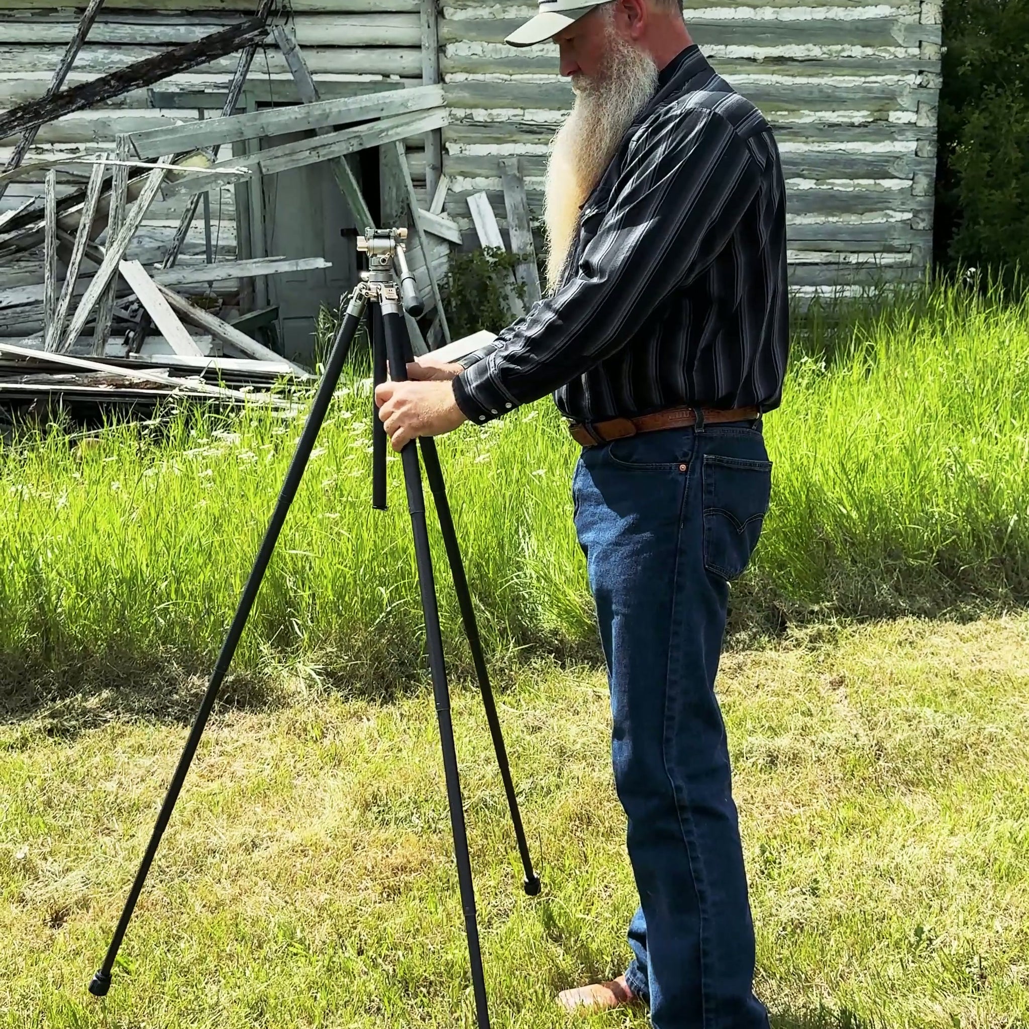 A man with a long white beard wearing a black and white striped shirt, blue jeans, and a baseball cap is adjusting a black tripod outdoors on a grassy lawn in front of a rustic wooden building.