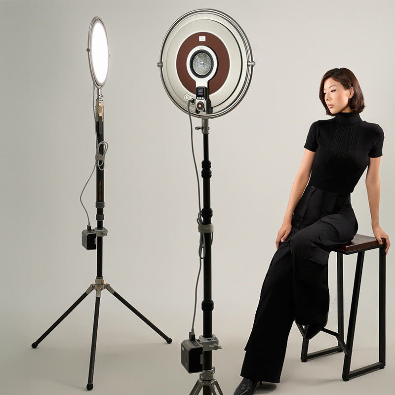 Harlowe Sol 100W round LED panel light with silver frame, illuminated front and back, with model on stool.